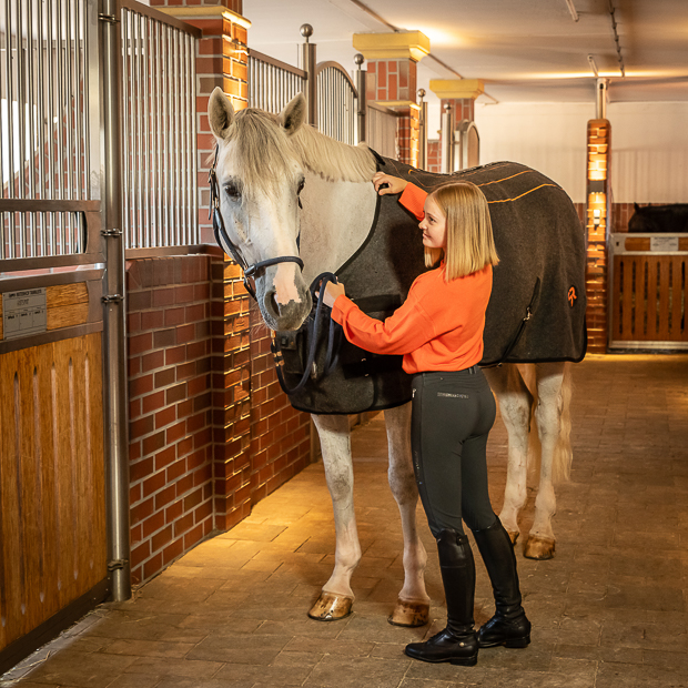 pferd mit grauer heizdecke von sunride mit seiner reiterin in der stallgasse