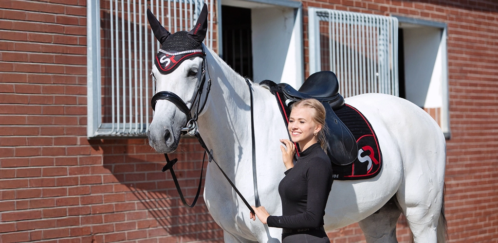 Eine Reiterin steht neben einem Pferd mit schwarz roter Schabracke und passender Fliegenhaube mit SR Logo vor einem Stall