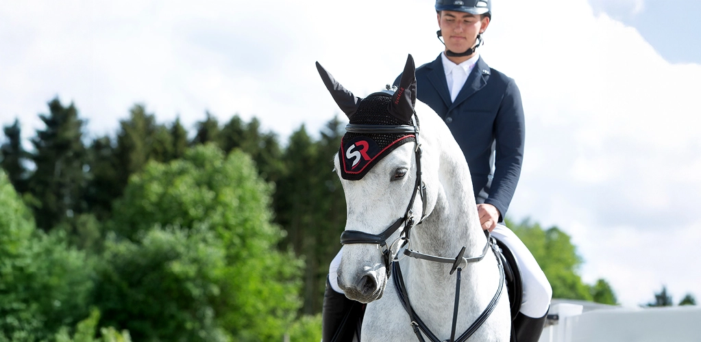 Fliegenhaube Ein Pferd mit roter Fliegenhaube und Reiter im Hintergrund sind grüne Bäume