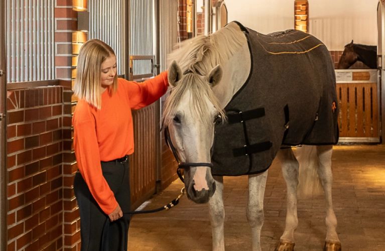 Heizdecke Eine Reiterin steht in der Stallgasse neben einem Pferd das eine Heizdecke trägt und streichelt es